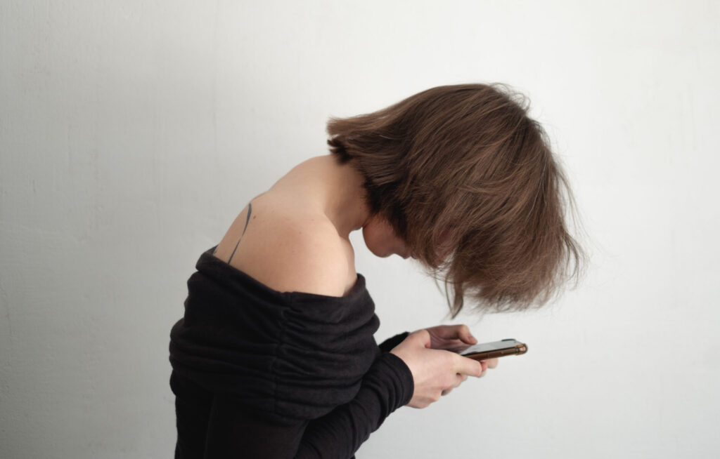 Woman with forward head posture and rounded shoulders looking down at phone, showing visible dowager’s hump