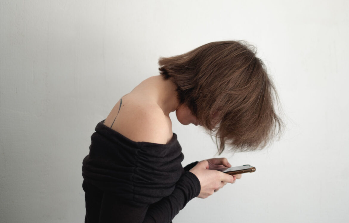 Woman with forward head posture and rounded shoulders looking down at phone, showing visible dowager’s hump